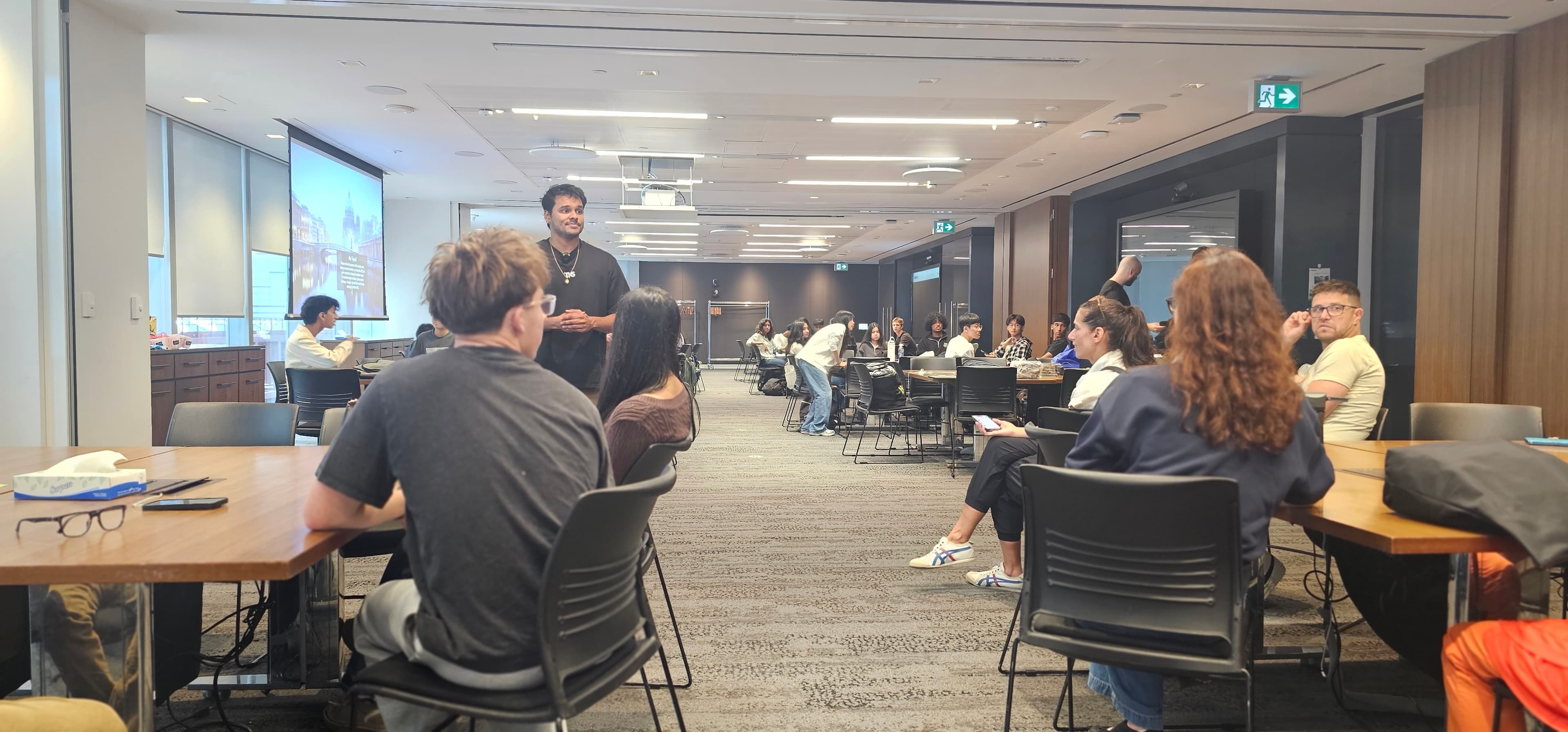 A man presents to students seated at tables in a large, modern conference room.