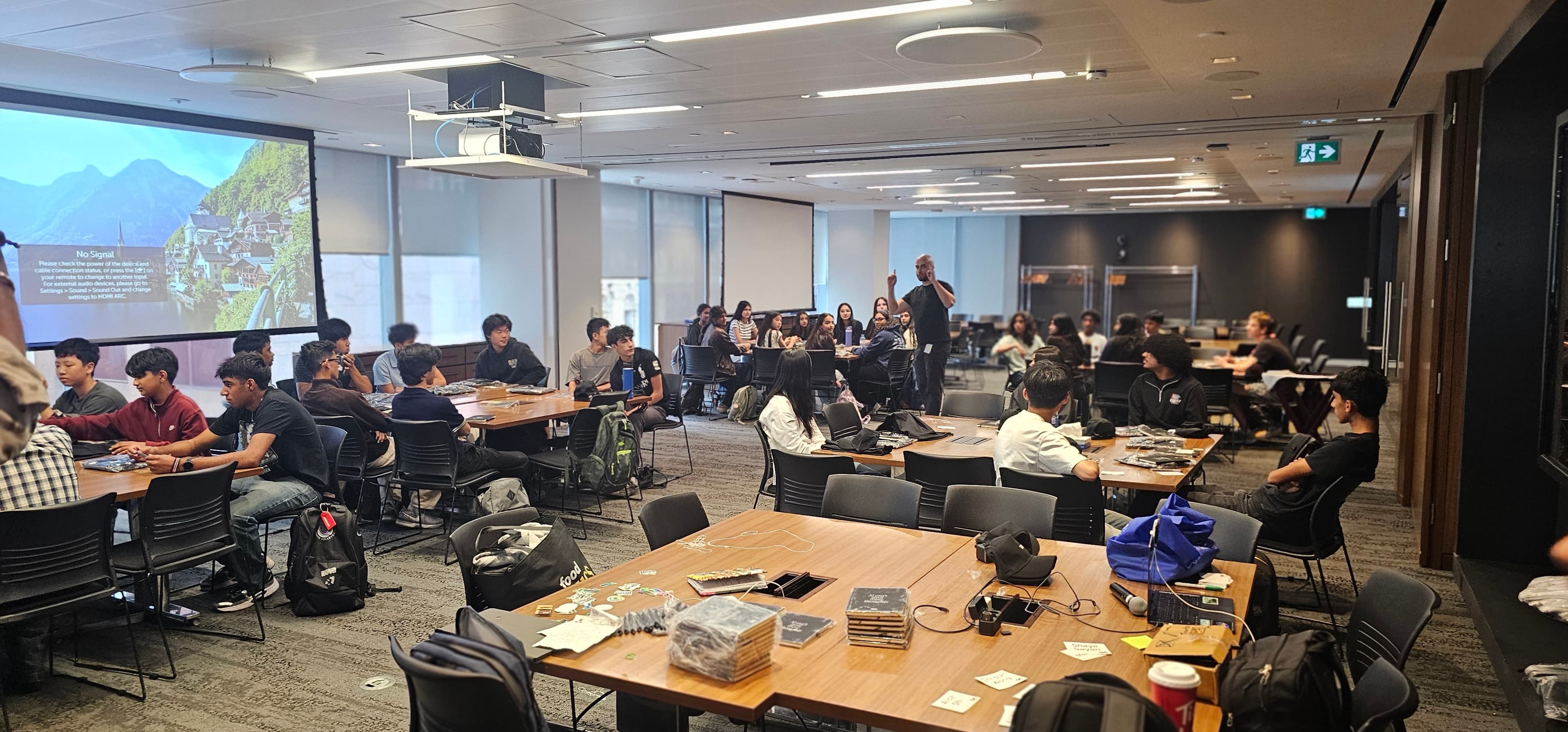 Students sit at tables in a large conference room while a man gives a presentation.