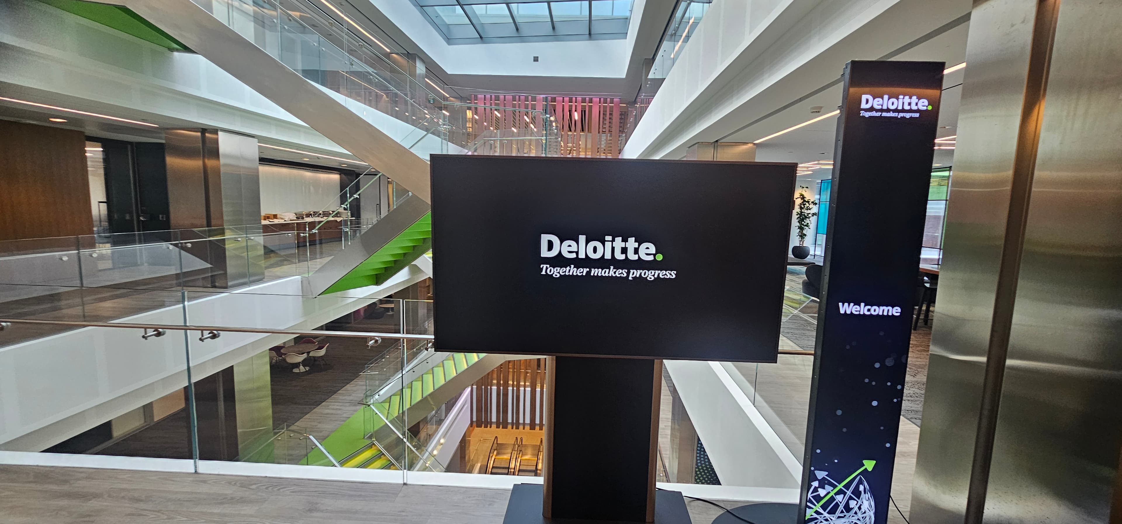 Deloitte logo on a digital screen in a modern, multi-level office atrium with glass railings.