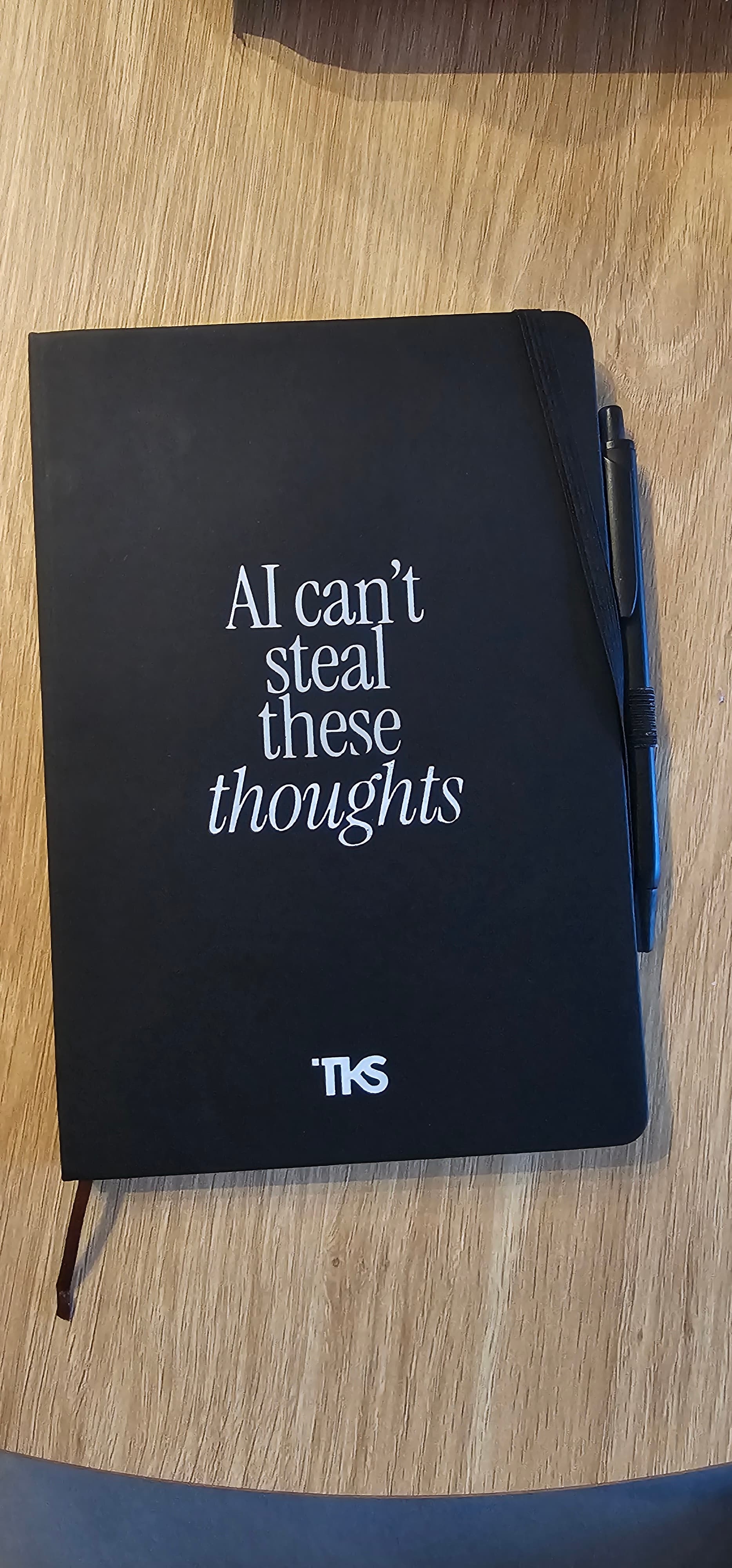 Black notebook on wood reading "AI can't steal these thoughts" with a pen attached.