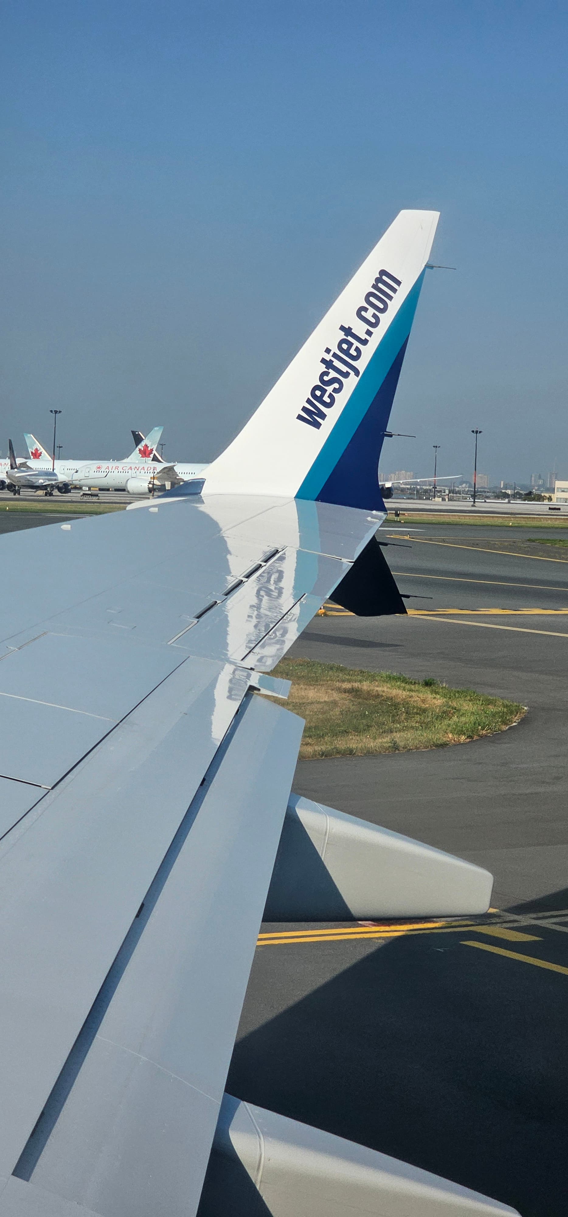 WestJet airplane winglet with website logo overlooking an airport tarmac and clear blue sky.