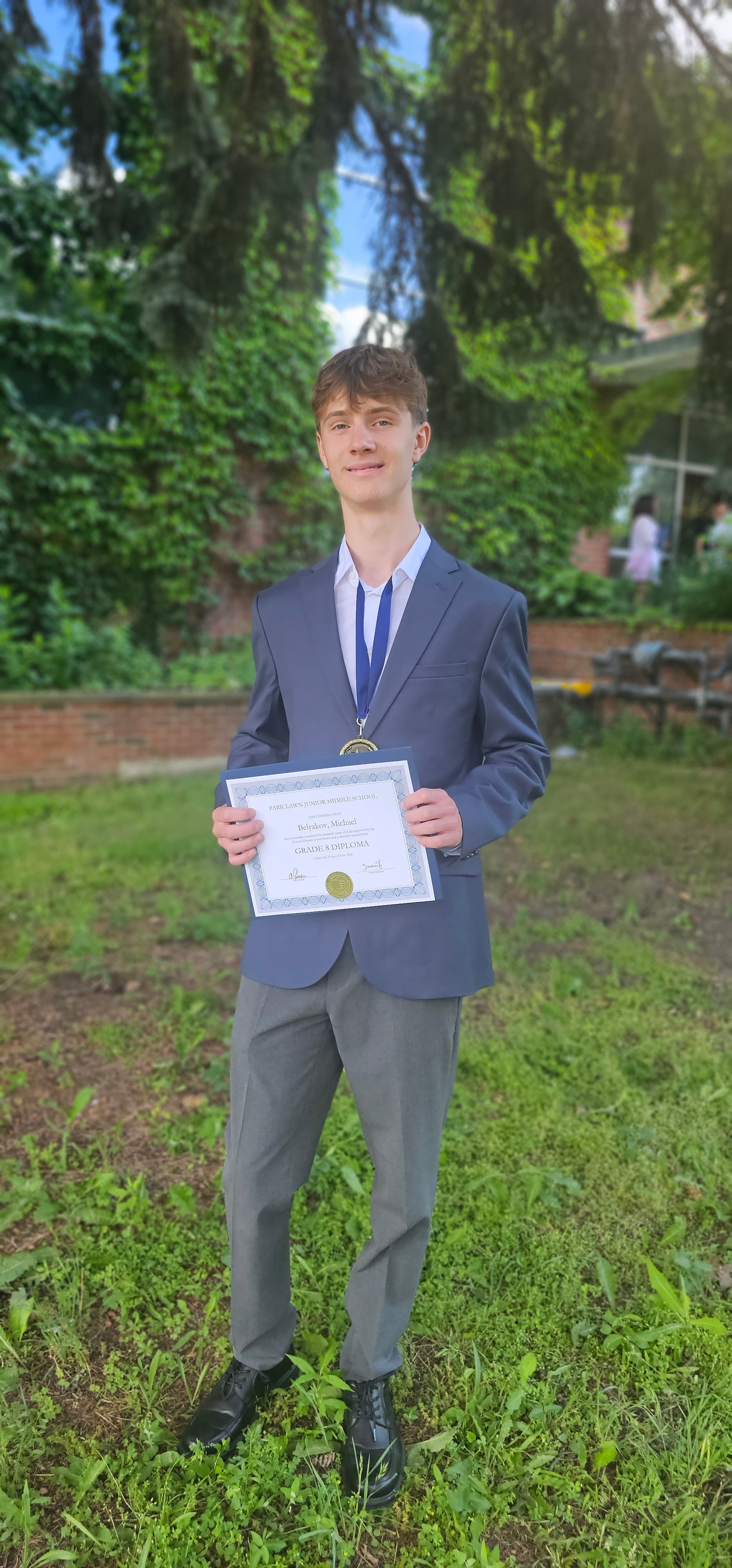 Young man in a blue blazer holding a Grade 8 diploma and wearing a medal.