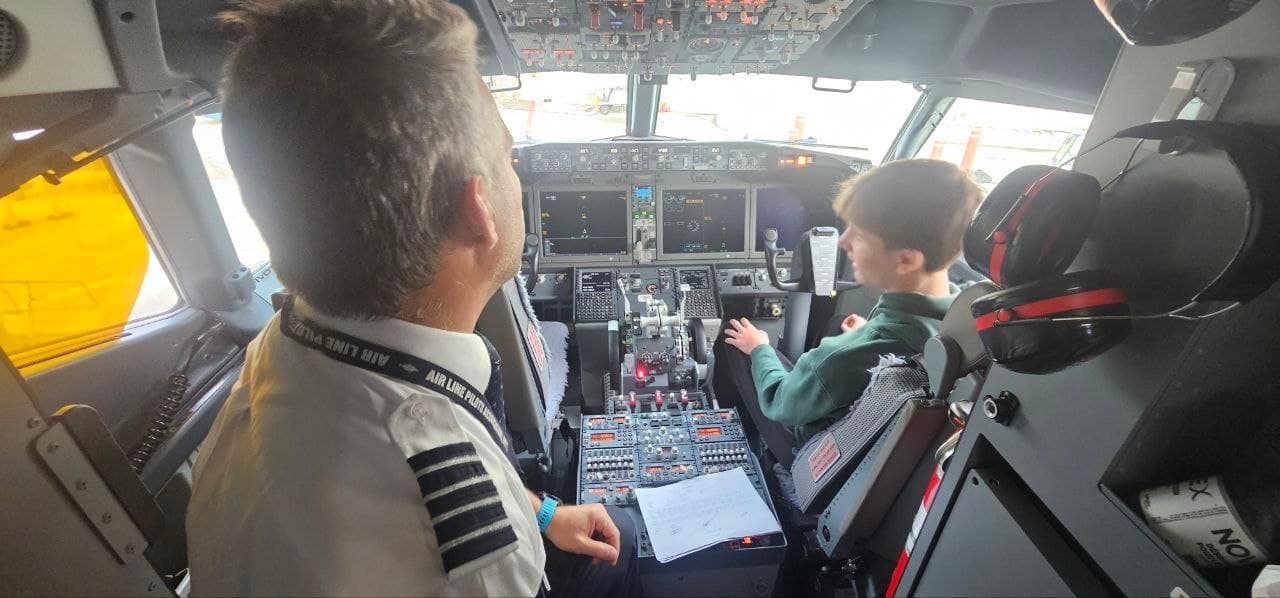 Pilot in uniform and young boy sit in a cockpit with complex flight controls.