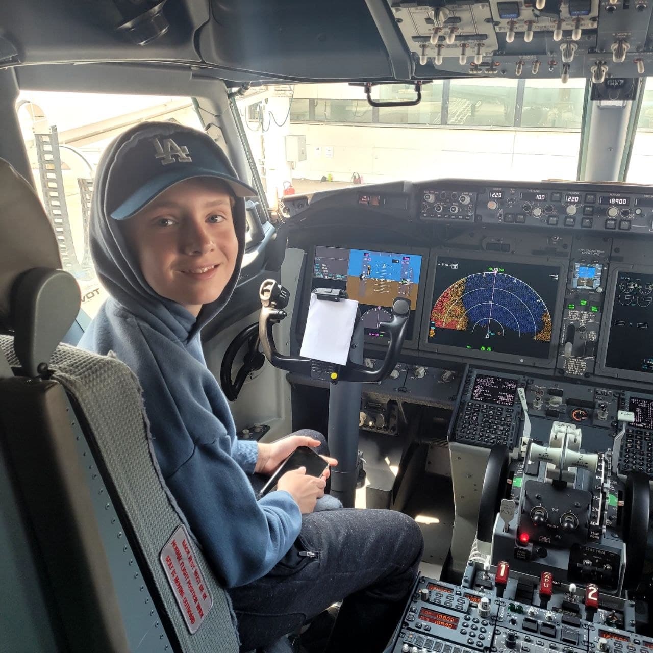 Smiling boy in a blue hoodie and LA cap sits in an airplane cockpit.
