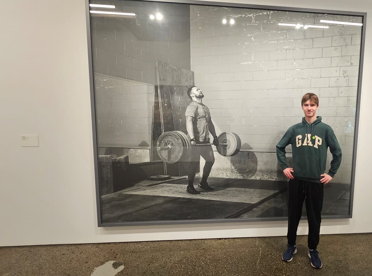 Young man in green hoodie standing next to a large black-and-white weightlifting photograph.