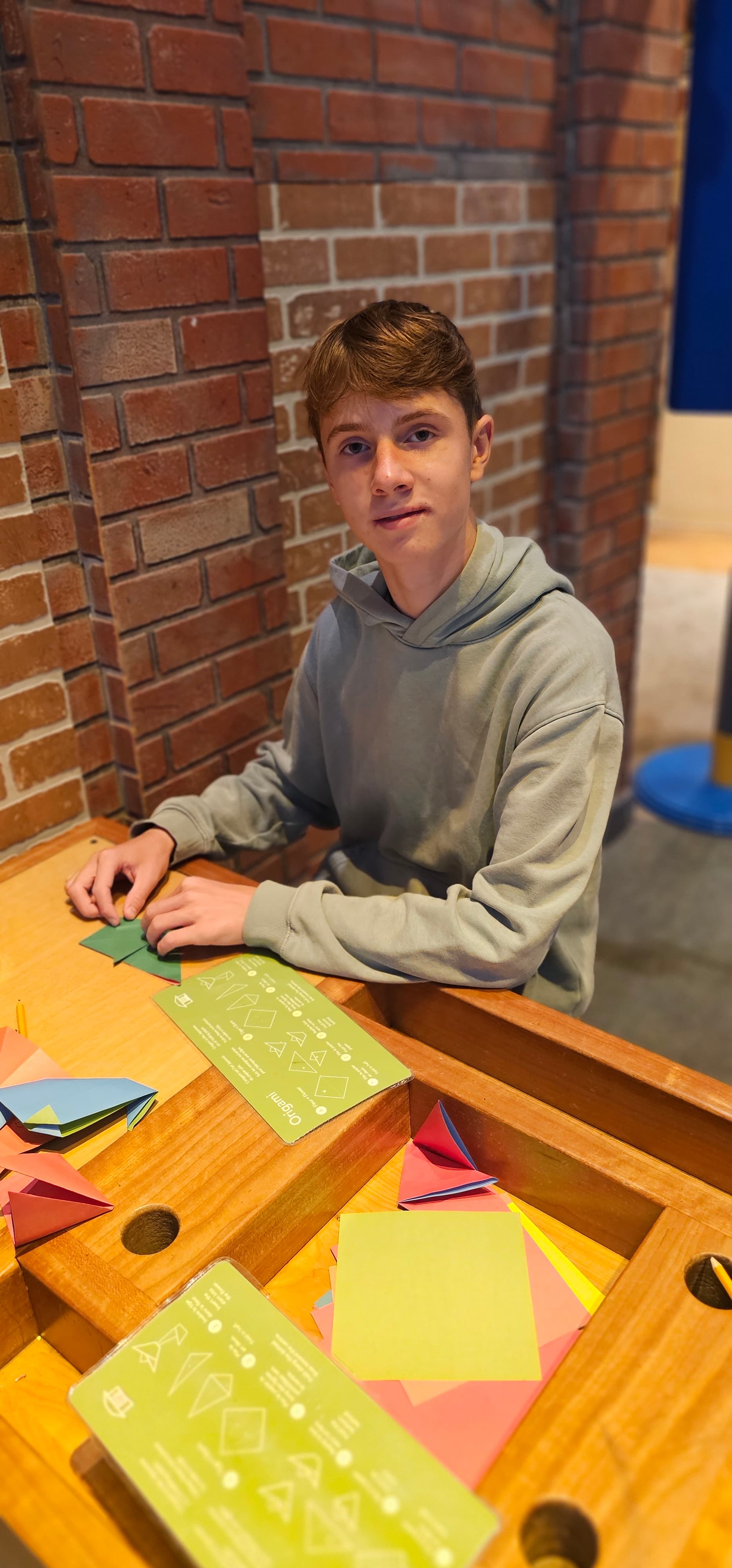 Young man in a grey hoodie folding green origami paper at a wooden table.