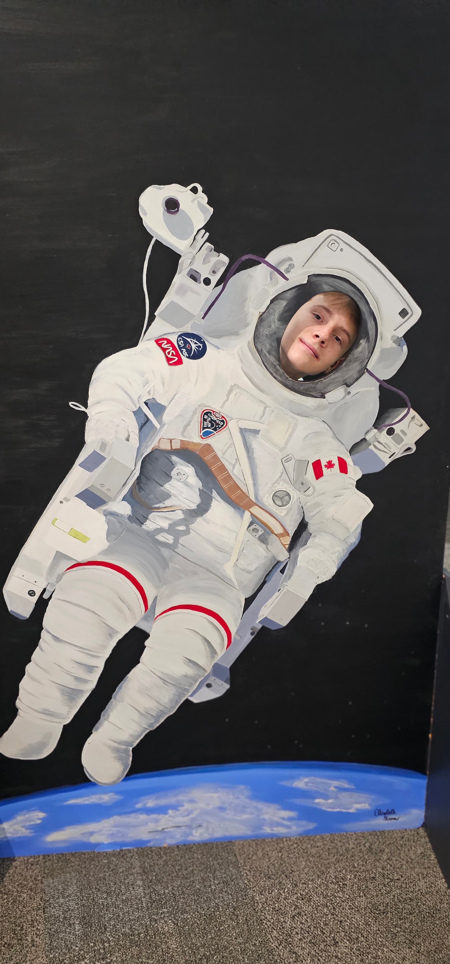 Young man's face in a painted astronaut suit cutout with NASA and Canadian flag patches.