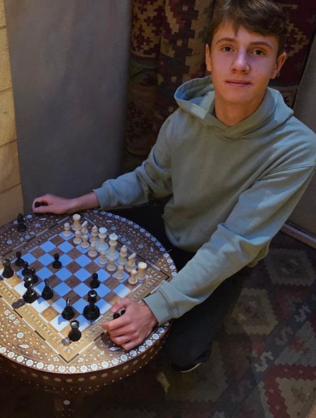 Young man in a green hoodie sits behind an ornate, round wooden chessboard table.