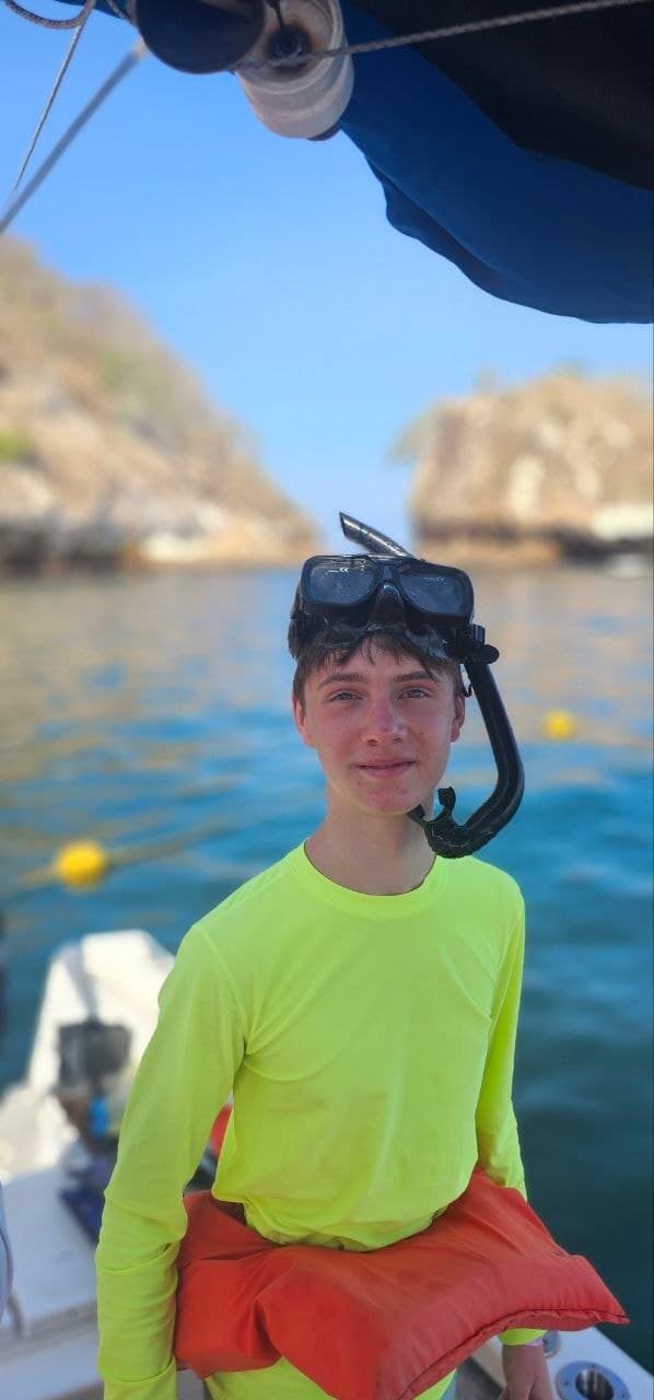 Boy in neon yellow shirt and snorkel mask on a boat near rocky tropical cliffs.