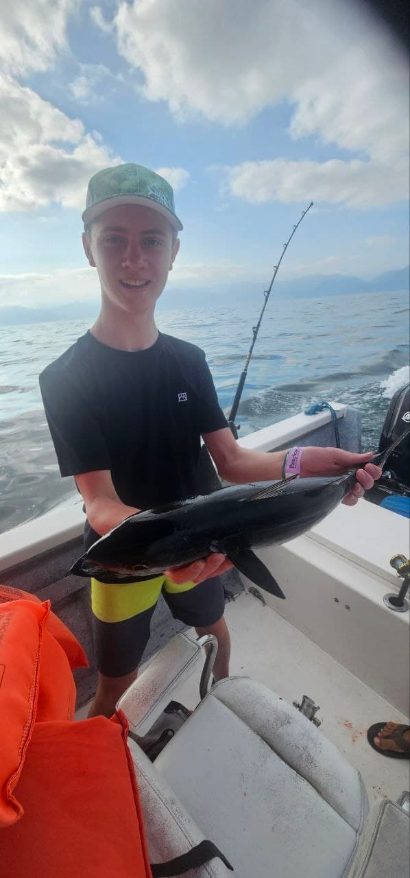 Smiling young man on a boat holding a dark tuna against a mountain backdrop.