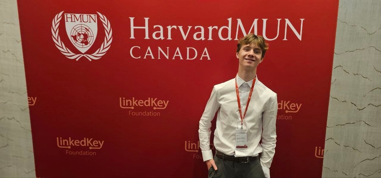 Smiling young man in a white shirt posing before a red HarvardMUN Canada event backdrop.