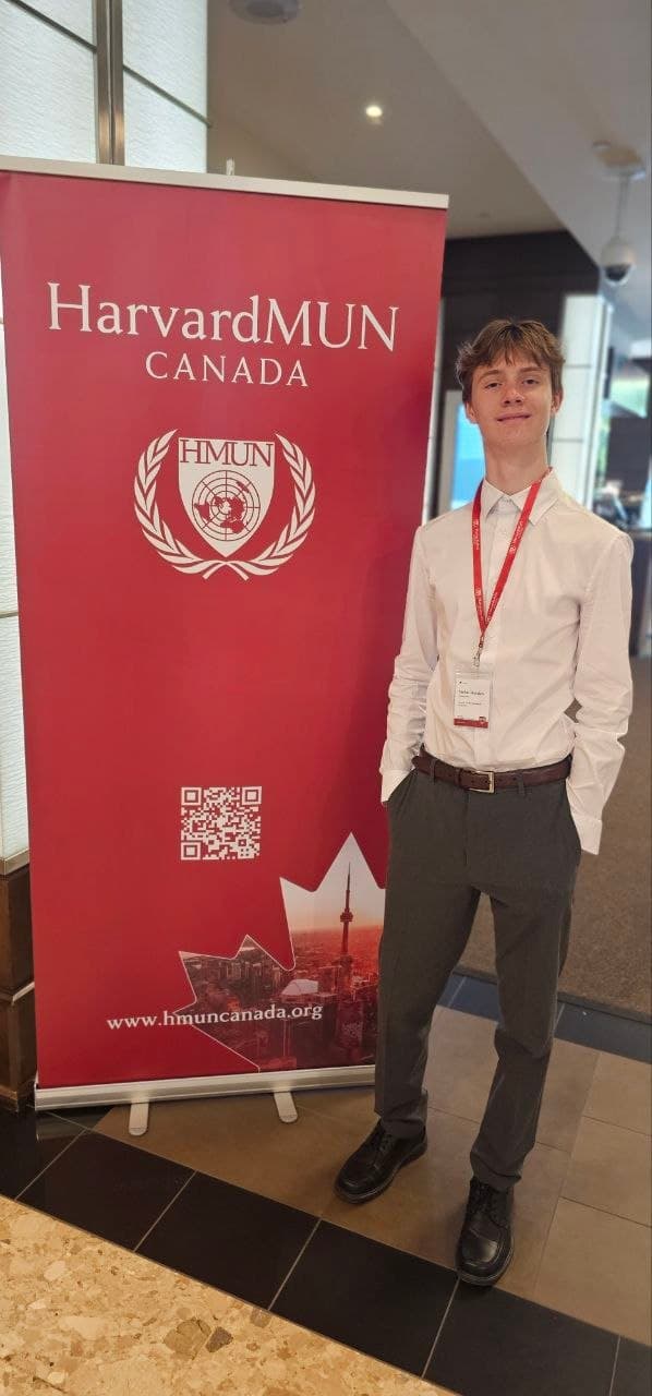 Smiling young man in formal wear stands beside a red HarvardMUN Canada banner.